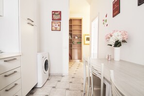 A kitchen with a washing machine, a dining area with chairs, and a hallway leading to a room with a bookshelf. - Alessia's Flat - Breno 1 (Milan)