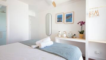 Una cama bien hecha con una manta azul y almohadas blancas, una mesita de noche con una planta y un ventilador de techo.