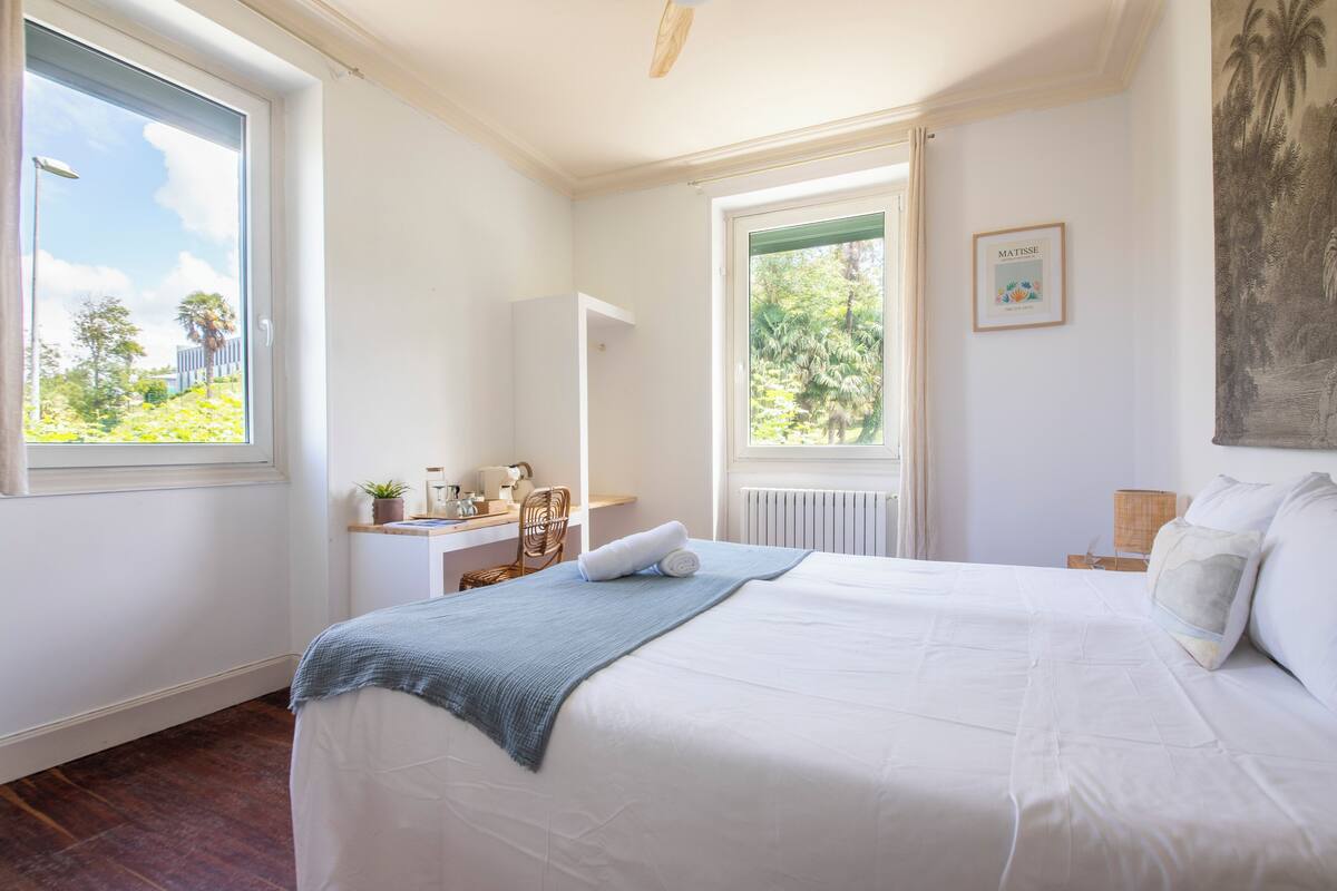 A bedroom with a large bed, a desk, and a window with a view of greenery.