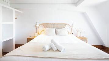 A neatly made bed with white linens and two rolled towels, a wooden headboard, bedside tables with lamps, and a white wall.