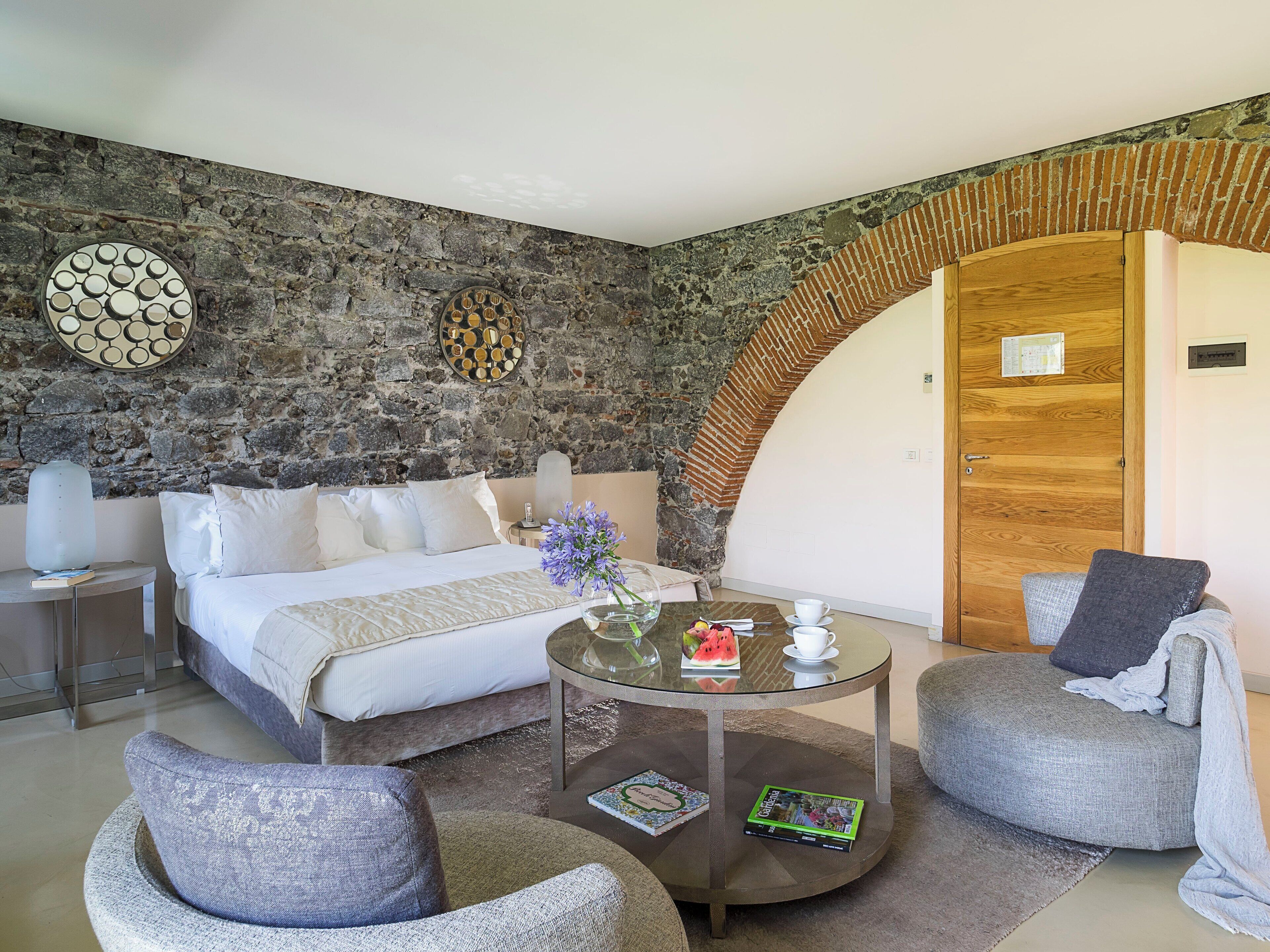 Une chambre avec un mur en pierre, une porte en bois, un lit, une table basse ronde en verre, deux fauteuils et une table de chevet avec une lampe.