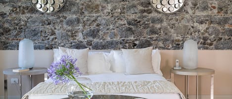 A modern bedroom with a stone wall, a bed with white bedding, two side tables with decorative mirrors, a glass coffee table with fruit and a cup, and a vase with purple flowers.