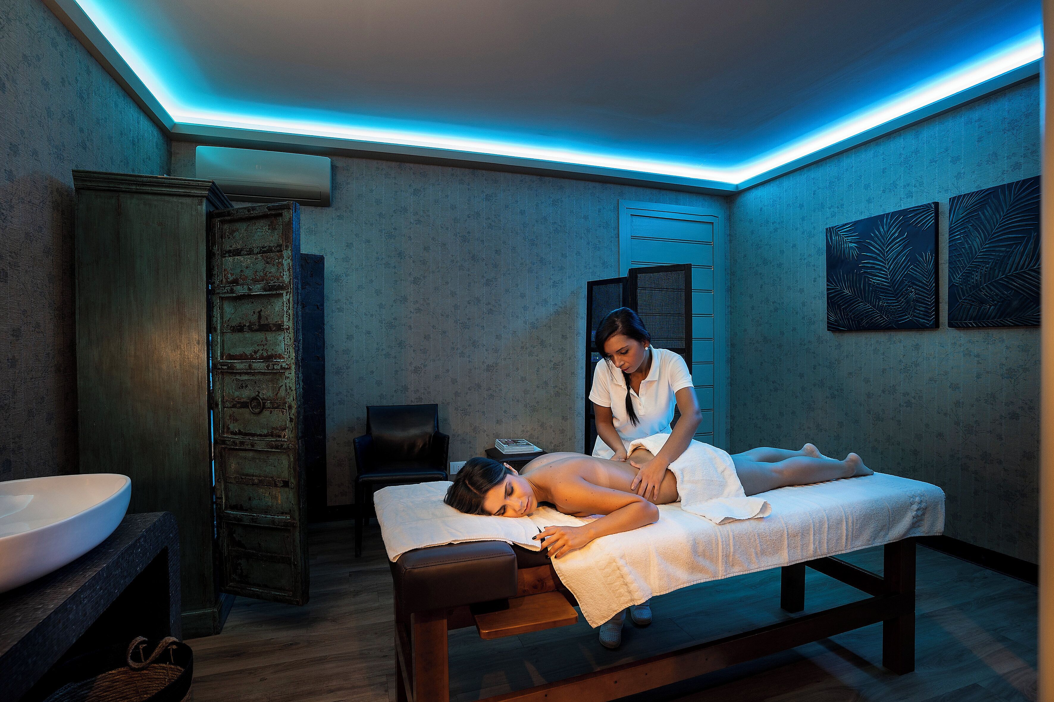 A massage therapy room with a wooden massage table, a white sink, and a towel-covered client.
