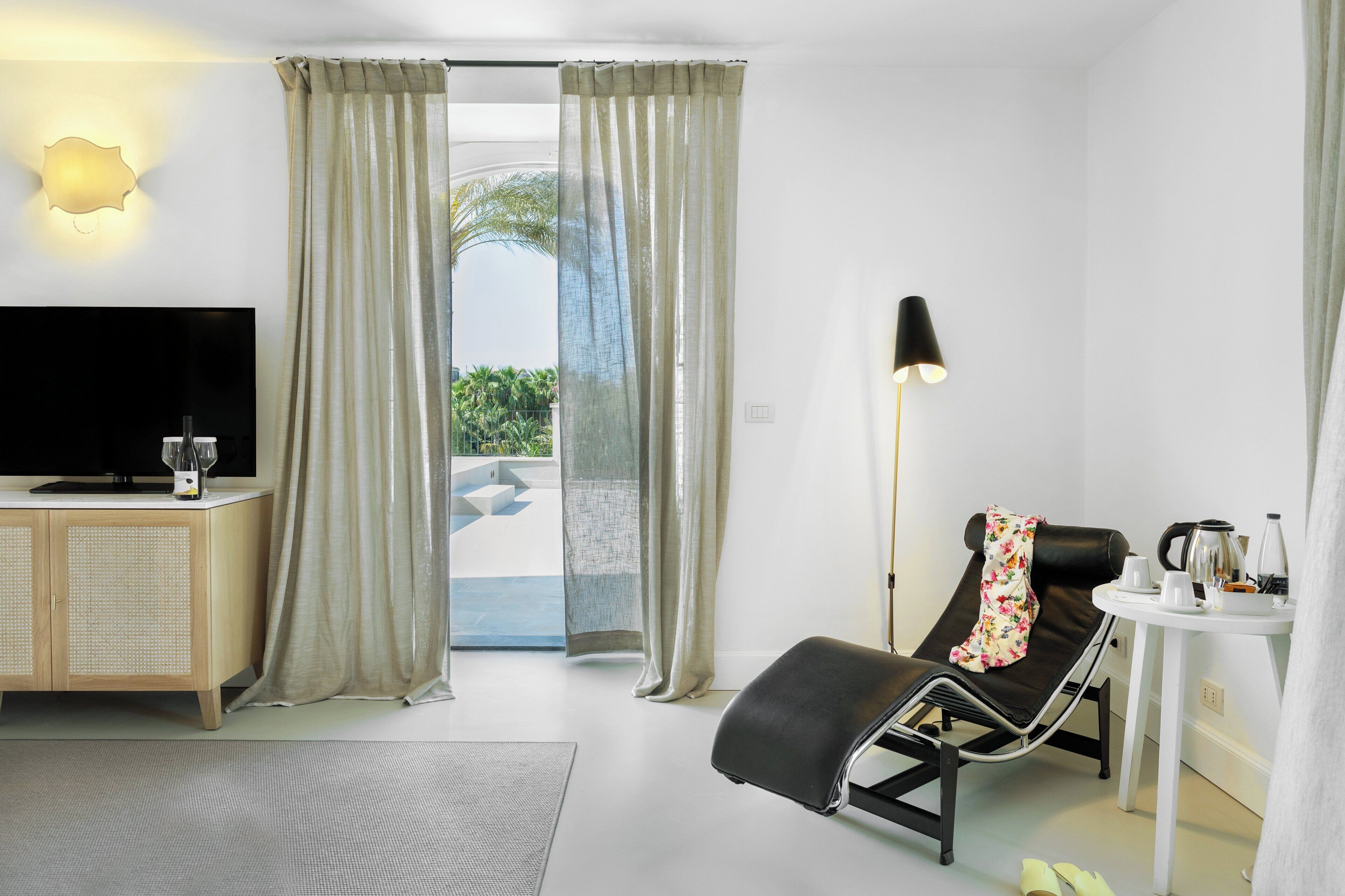 Un dormitorio de hotel moderno con televisión de pantalla plana, un sillón, una mesita de noche con lámpara y vistas al exterior.