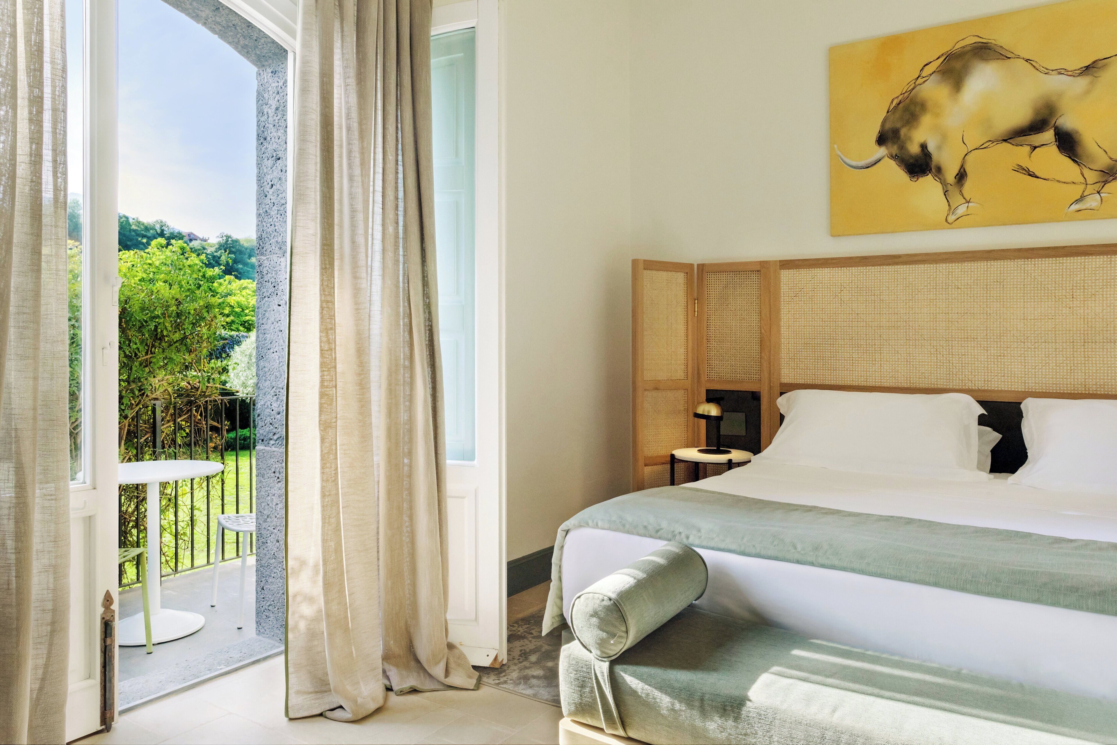 A hotel room with a bed, a view of greenery, and a painting of a bull on the wall.