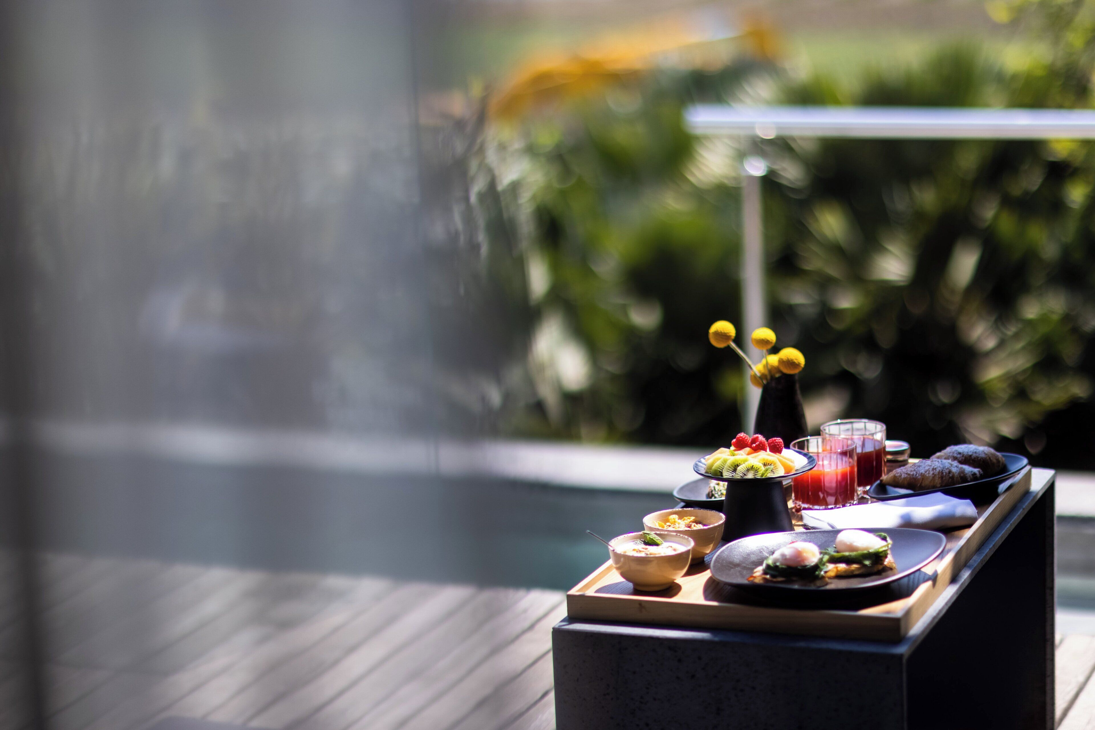 Un plateau de petit-déjeuner contenant divers aliments et boissons, posé sur une table en bois près d’une piscine.