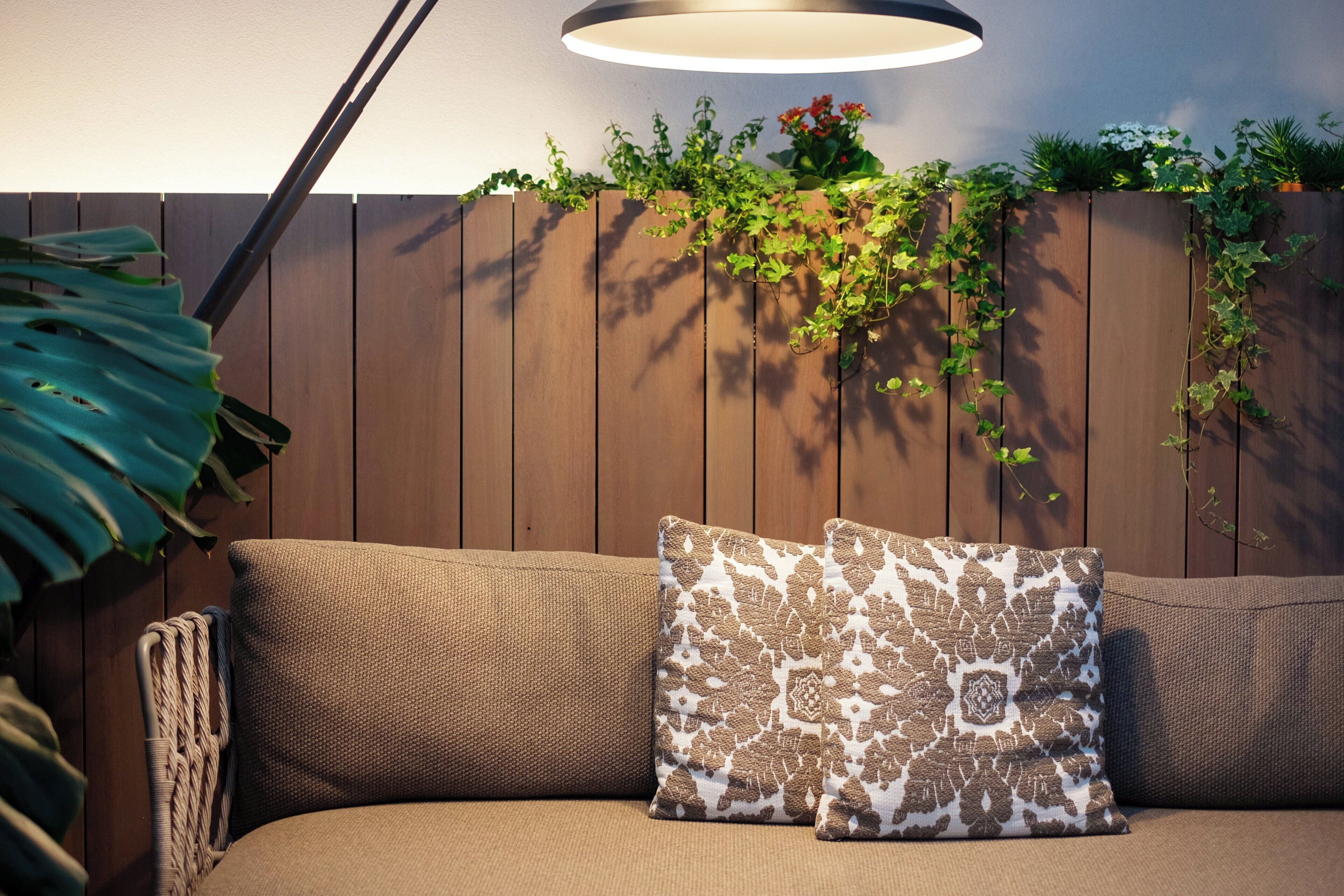 A wooden fence with climbing plants and flowers, a sofa with patterned cushions, and a hanging light fixture.