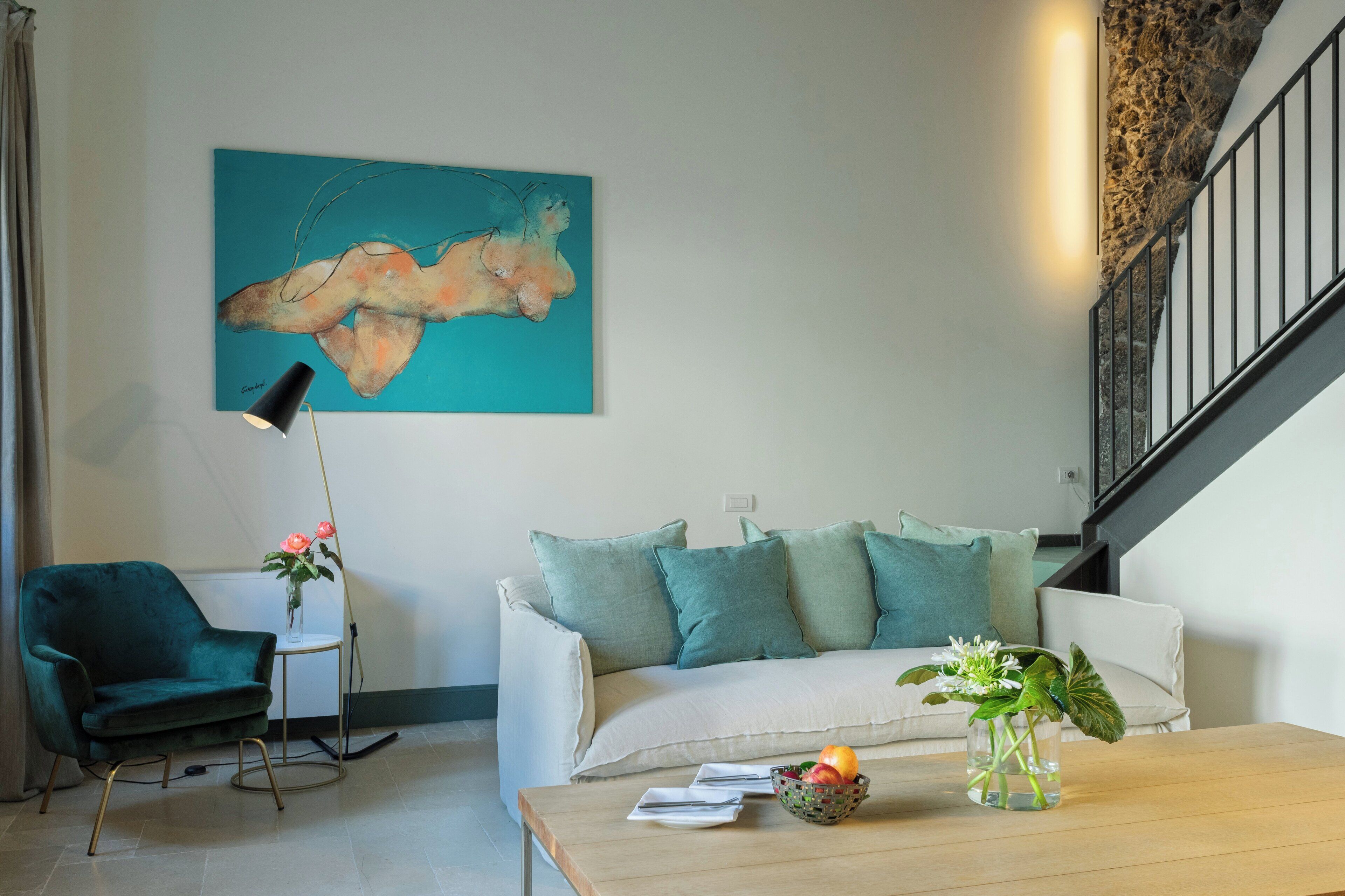 Una sala de estar moderna con un sofá color turquesa, un sillón verde, una mesa de centro con un florero y frutas, y un cuadro en la pared.