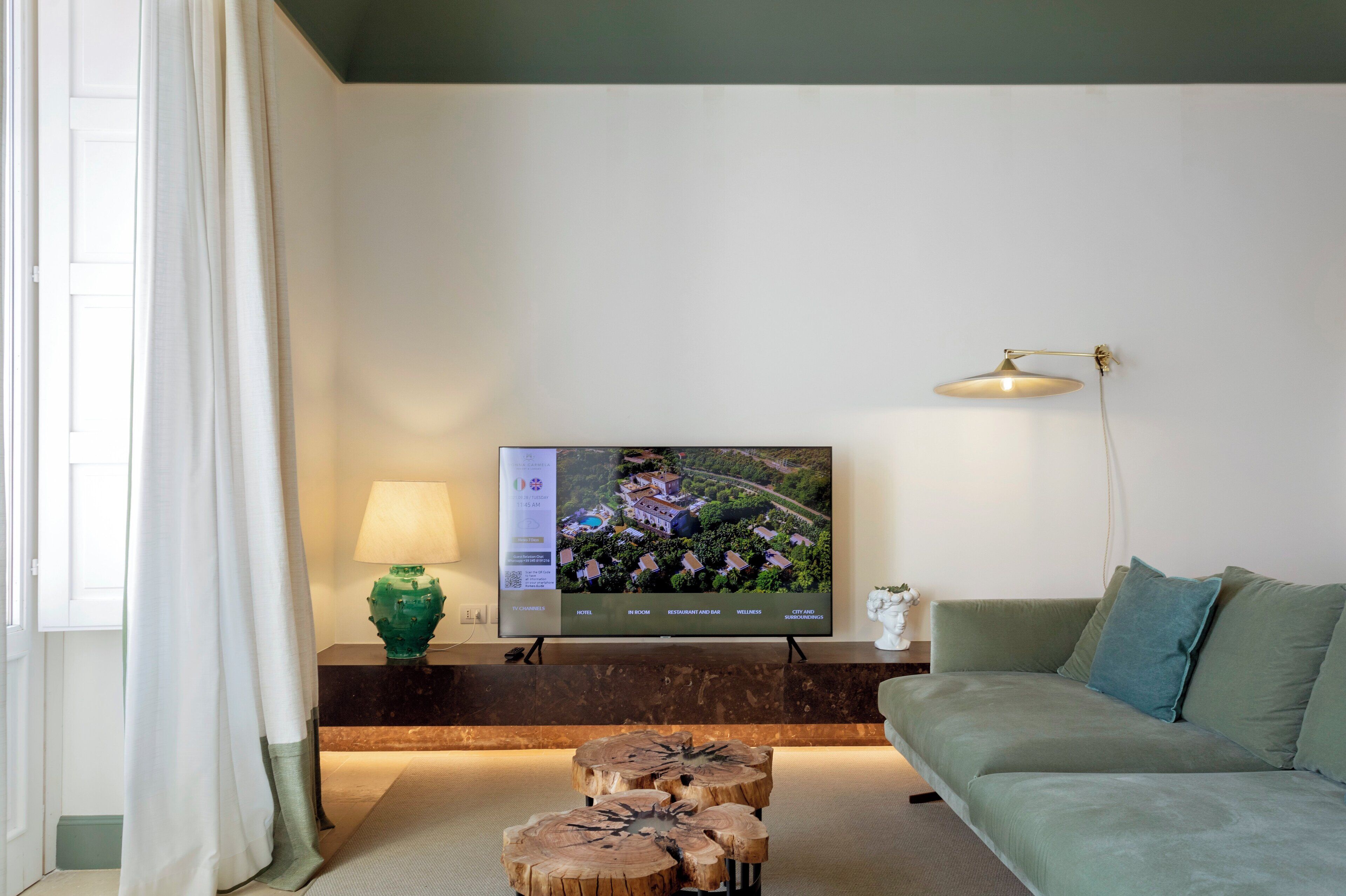 A modern living room with a flat-screen TV mounted on the wall, a green sofa, a wooden coffee table, a lamp, and a view of a property on the screen.