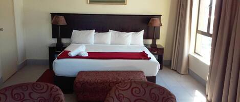 A hotel room with a large bed, two bedside tables, a red throw blanket, a painting on the wall, and a window with curtains.
