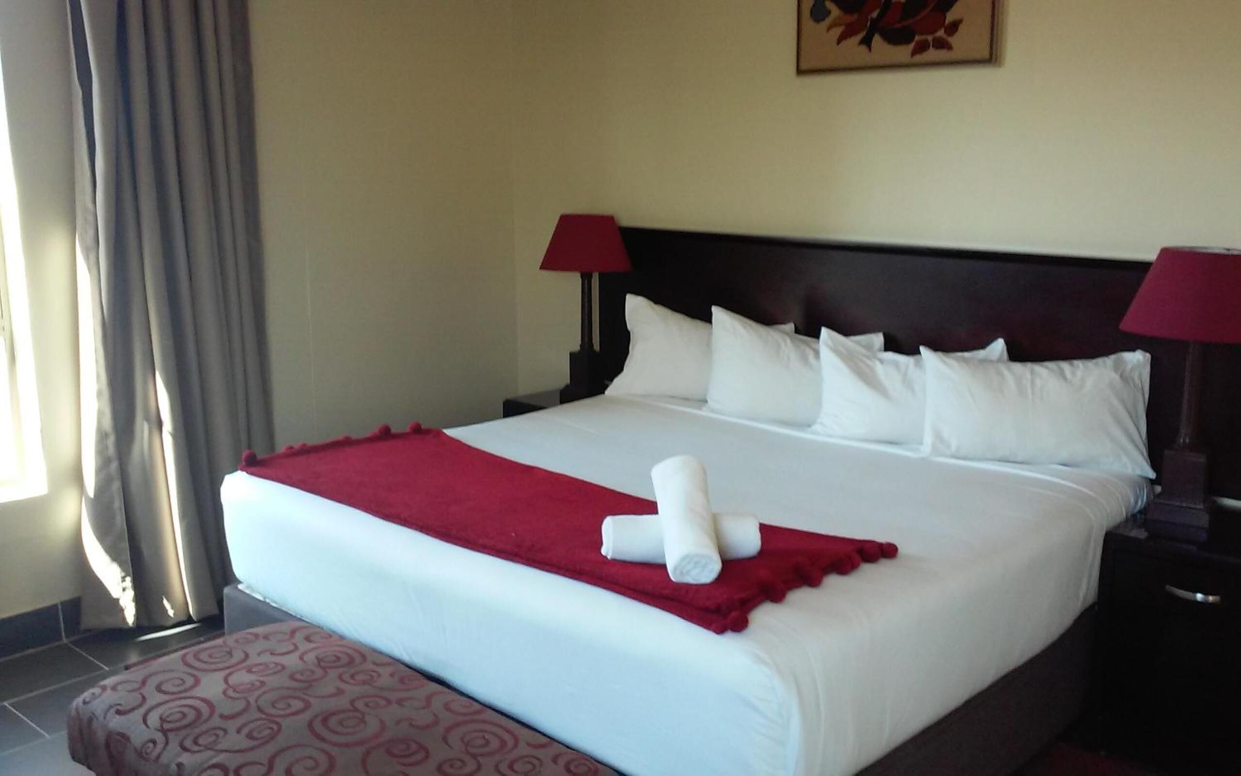 A neatly made bed with white linens and a red throw blanket, two red lamps on either side, and a painting on the wall.