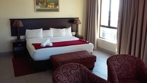 Una habitación de hotel con una cama grande, dos mesitas de noche, un cuadro en la pared, una ventana con cortinas y dos sillones estampados.