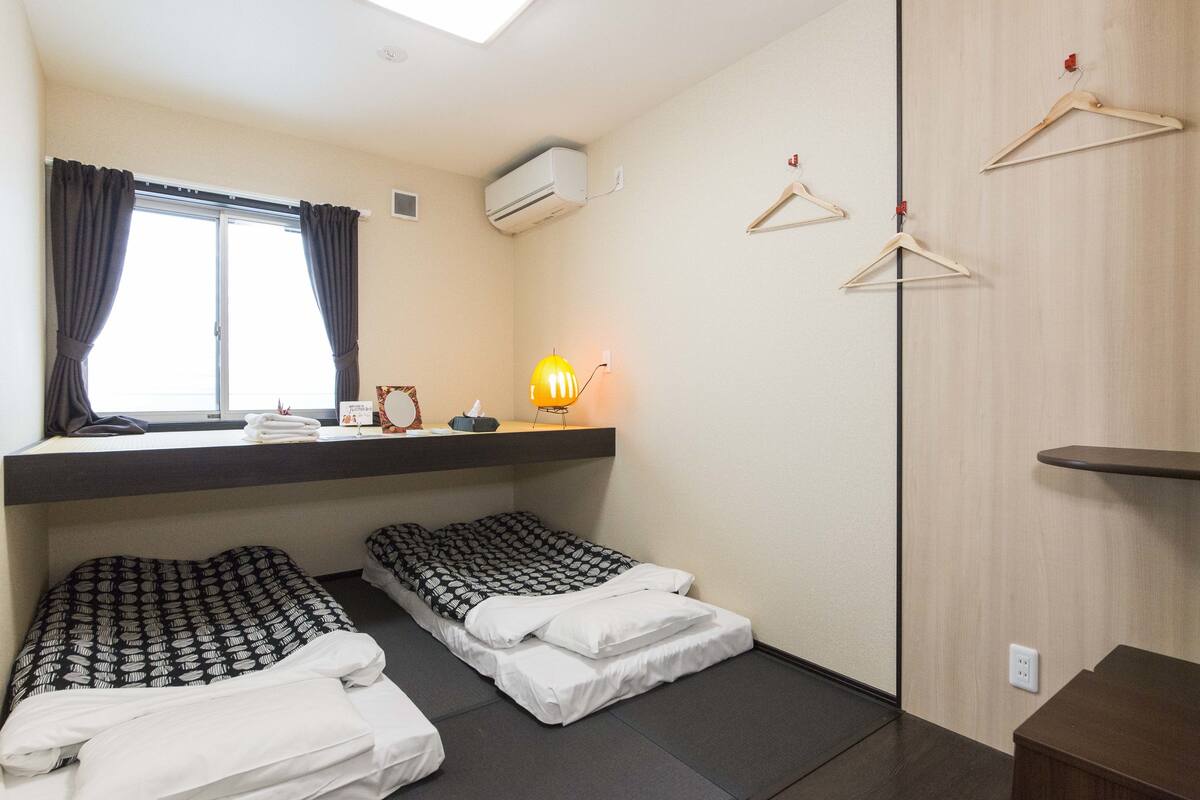 A compact room with two beds, a window with curtains, a wall-mounted air conditioner, a bedside lamp, and a wooden wardrobe with hangers.