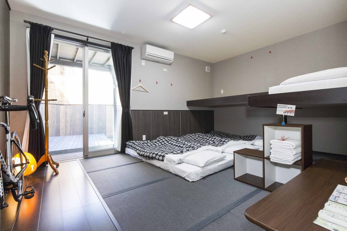 A hotel room with a bed, a desk, a chair, a bicycle, and a coat rack.