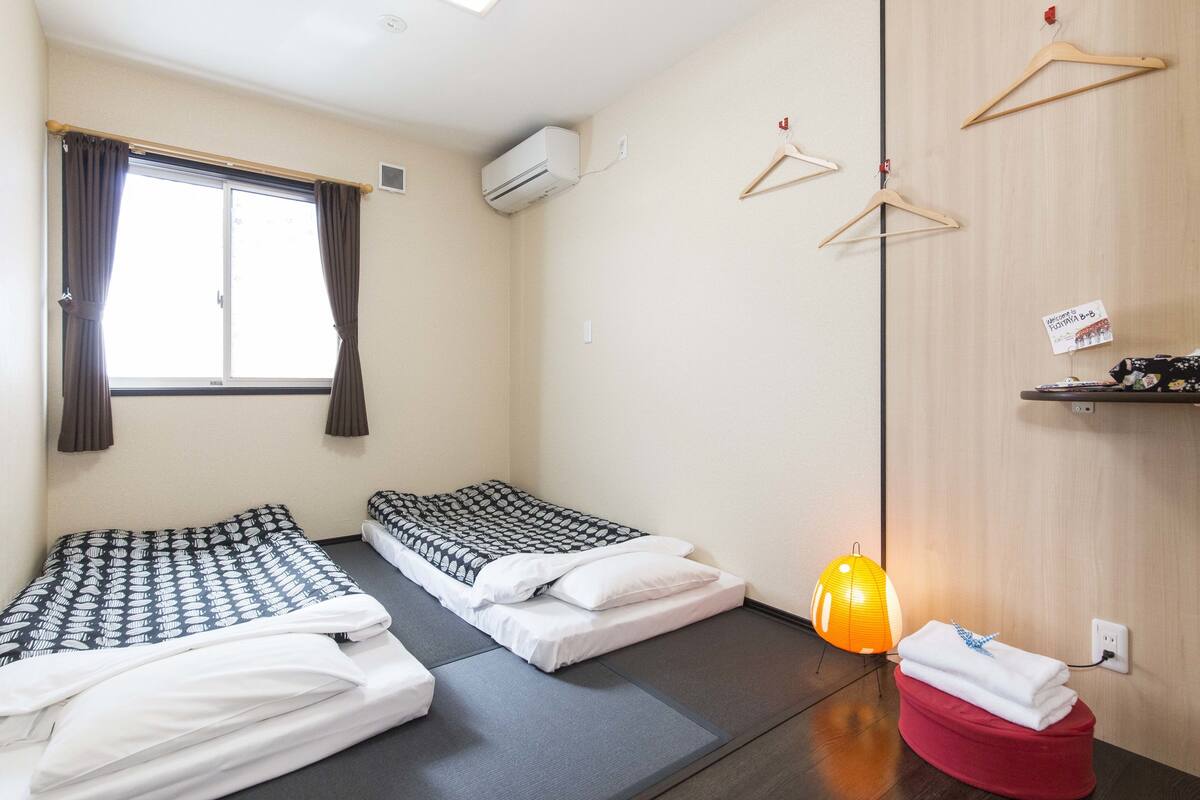 A room with two single beds, a red ottoman, a hanging light, and wooden shelves.