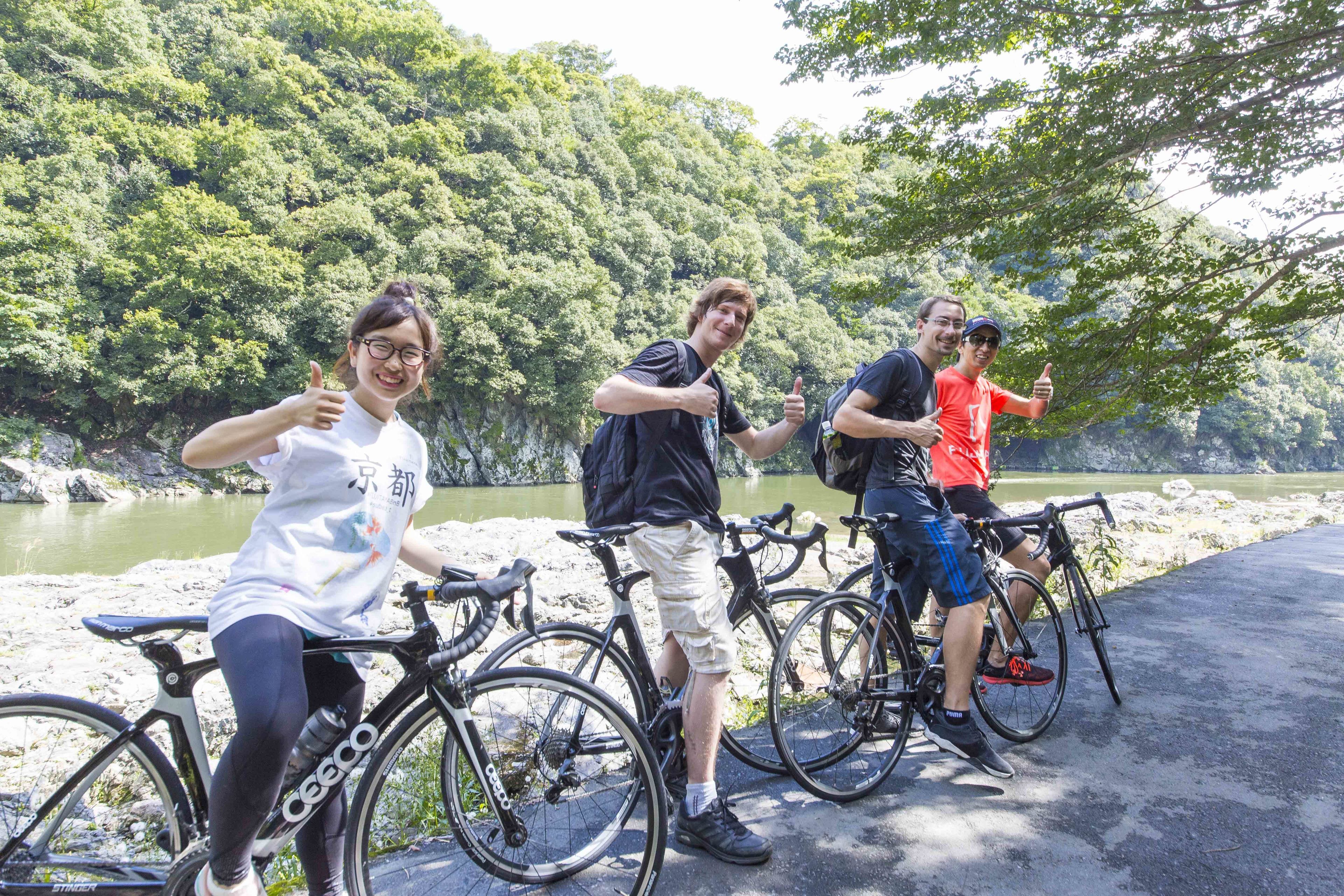 Un grupo de personas de pie junto a bicicletas cerca de un río, con una ladera boscosa al fondo.