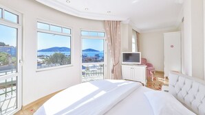 A bedroom with a bed, a TV, a wardrobe, and a balcony with a view of the sea and mountains. - Buyukada Princess Hotel (Adalar)