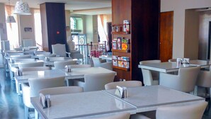A restaurant with white tables and chairs, a bookshelf with magazines, and a view of the street outside. - Buyukada Princess Hotel (Adalar)