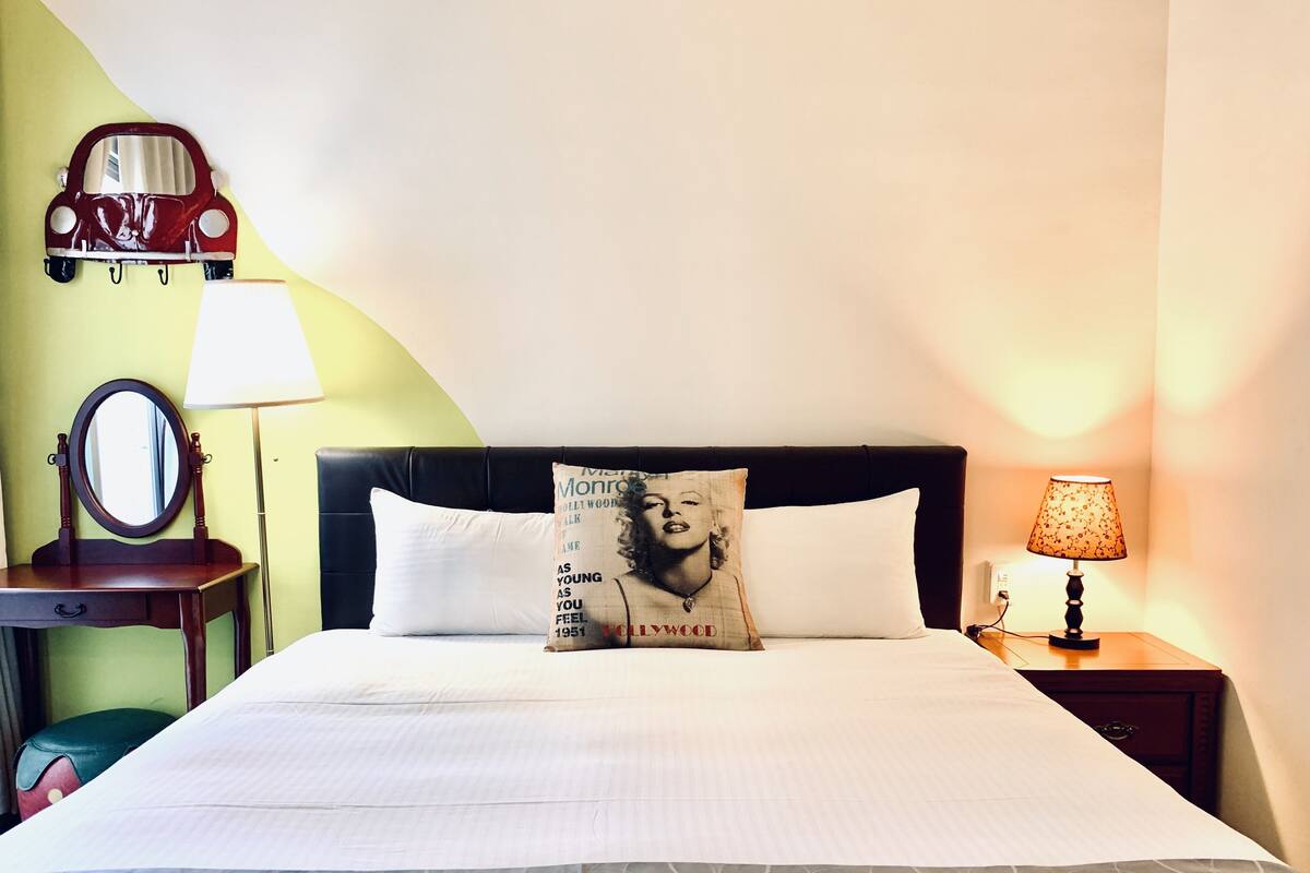 A neatly made bed with a pillow featuring a vintage design, a bedside lamp, and a mirror on a bedside table.