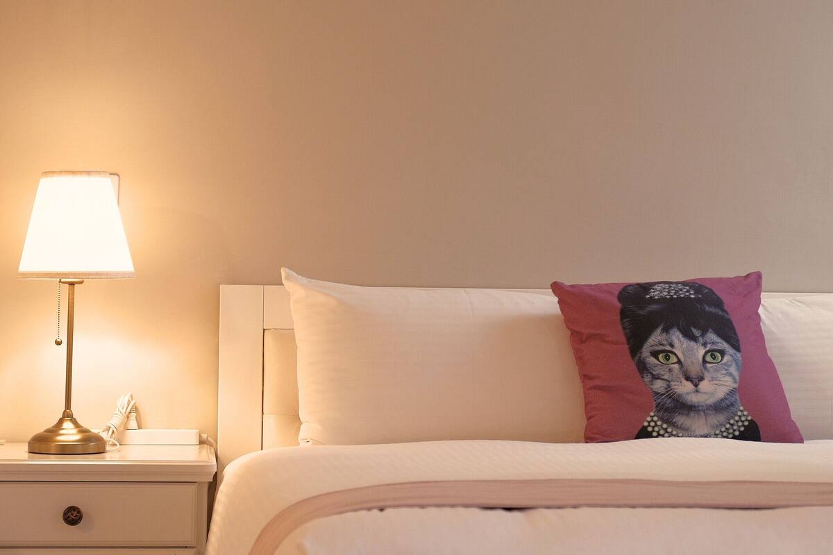 A neatly made bed with a pillow featuring a cat design, a bedside lamp, and a nightstand with a drawer.