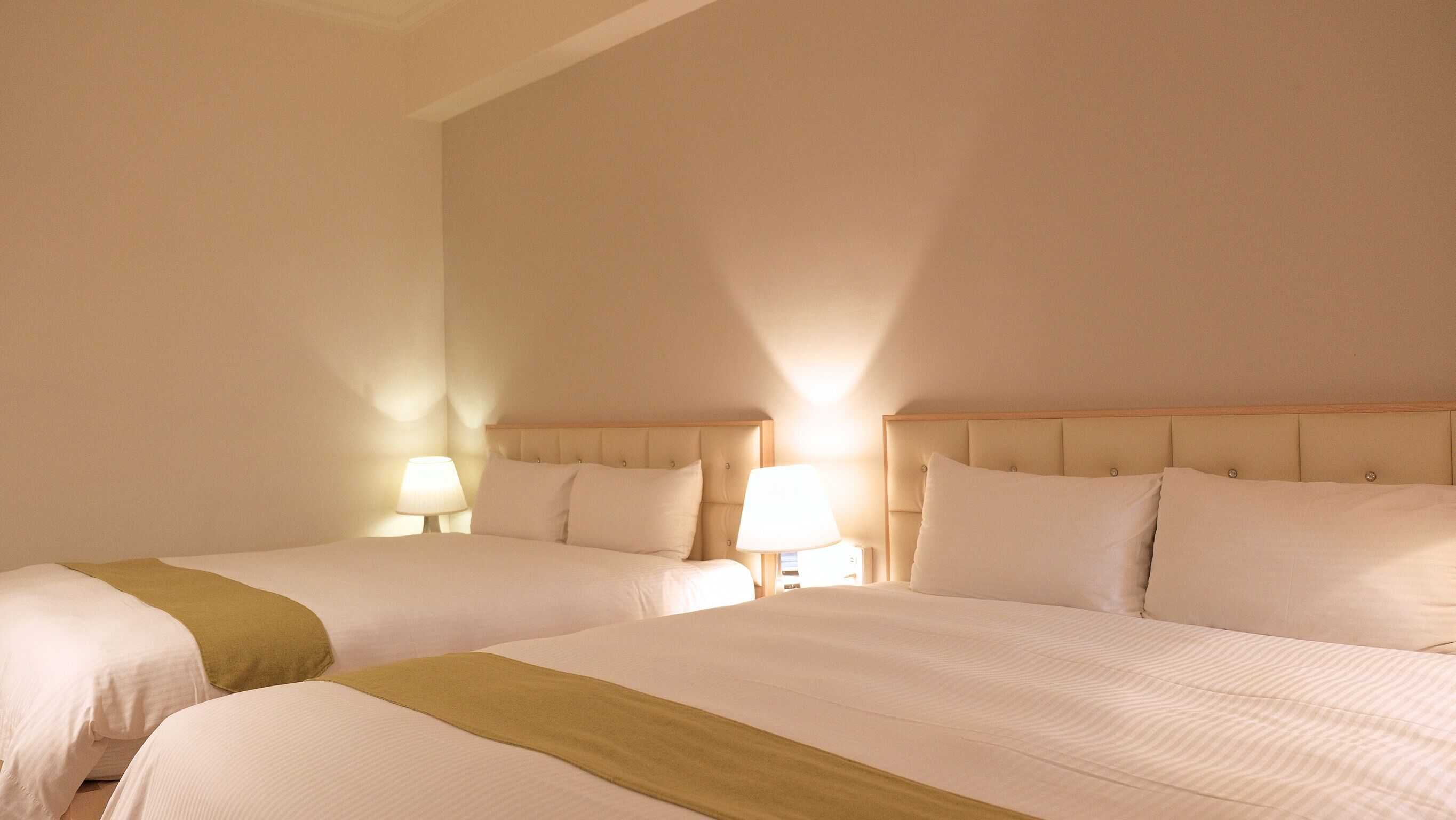 A hotel room with two beds, each with a headboard, bedside lamps, and a brown throw blanket.