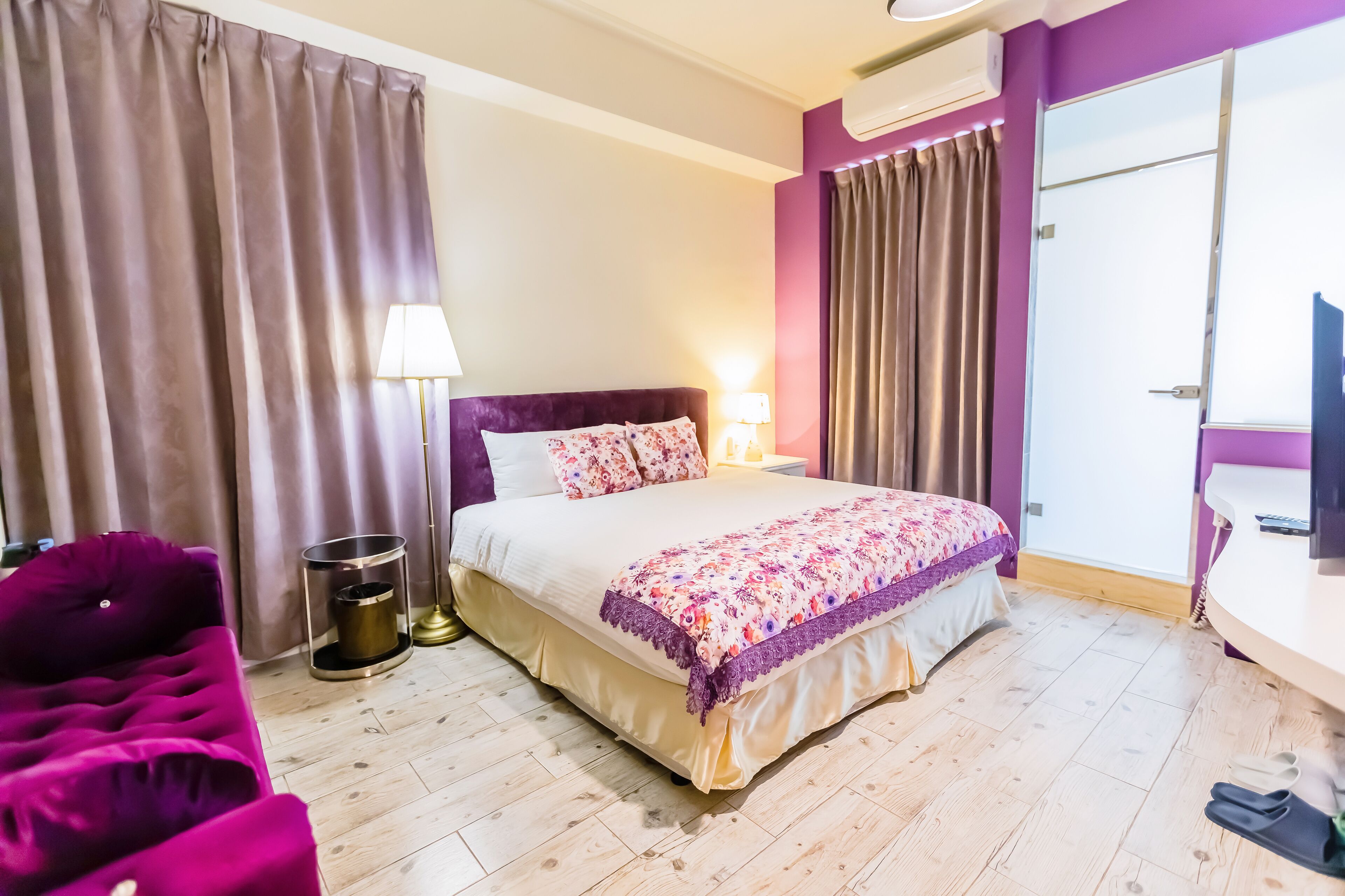 A hotel room with a bed, a purple armchair, a bedside table with a lamp, and a television.