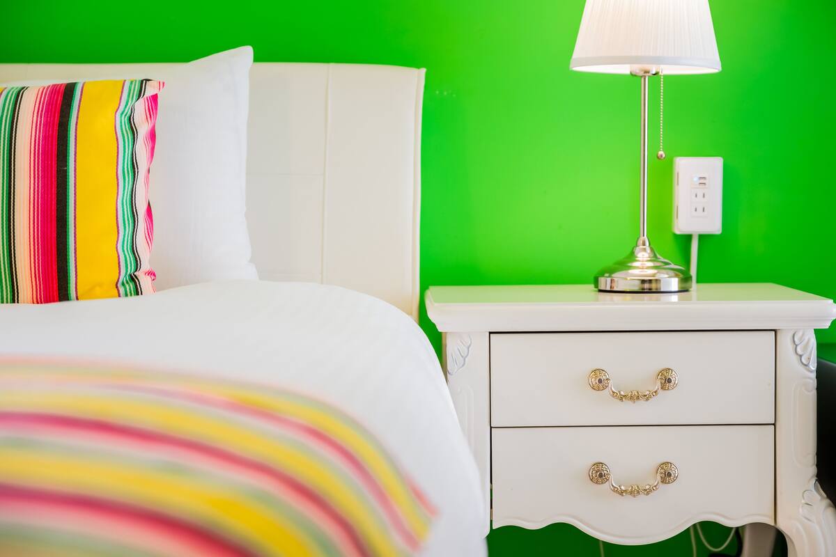 A bed with a colorful striped pillow, a white nightstand with two drawers, and a lamp.