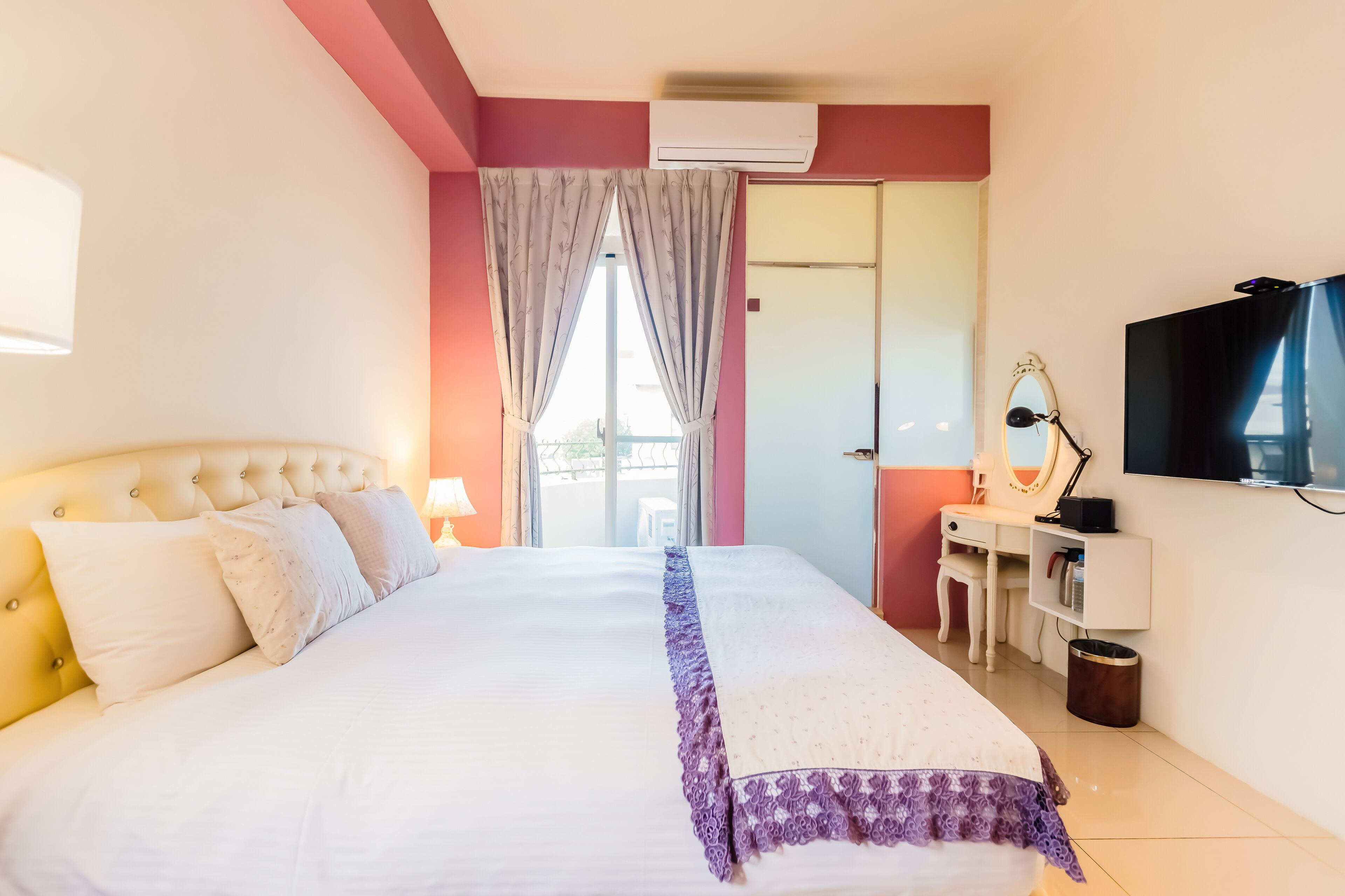 A hotel room with a large bed, a desk with a chair, a TV, and an air conditioning unit.