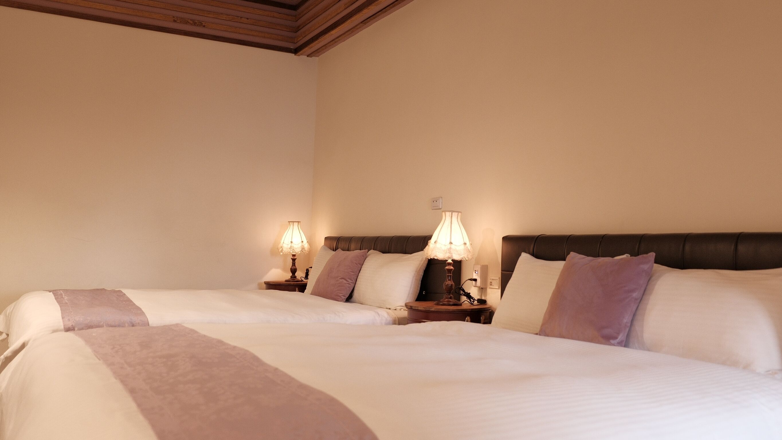 Habitación de hotel con dos camas, cada una con una mesita de noche y una lámpara. La habitación tiene techo de madera y paredes lisas.