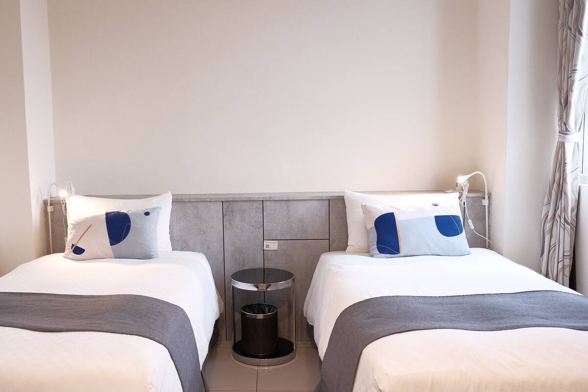 A hotel room with two single beds, a grey headboard, a waste bin, and a bedside table with a lamp.