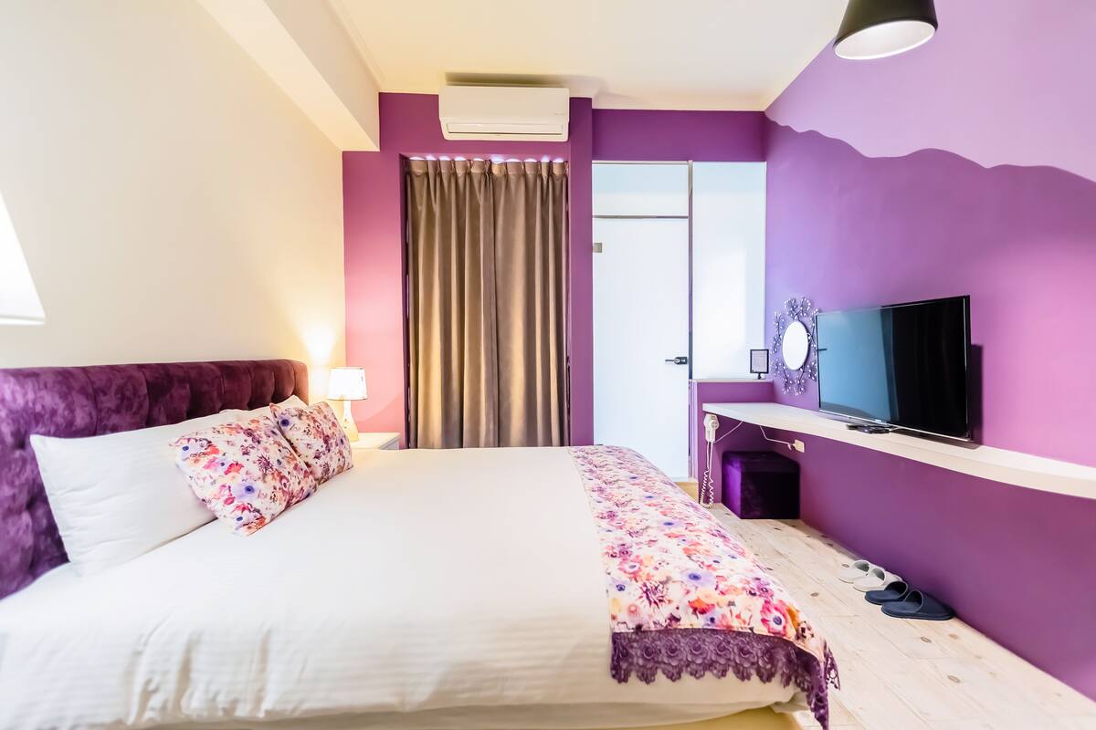 A bedroom with a bed, a TV, a desk, and a window with curtains.