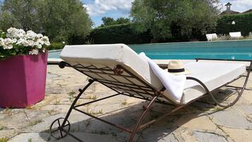 Una tumbona con sombrero y toalla, cerca de una piscina con solárium de piedra y vegetación.