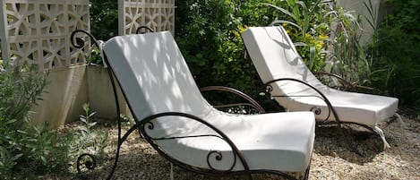 Zwei Liegestühle mit Metallgestellen und weißen Kissen, auf einem Kiesgrundstück mit dekorativem Zaun und Grünpflanzen im Hintergrund.