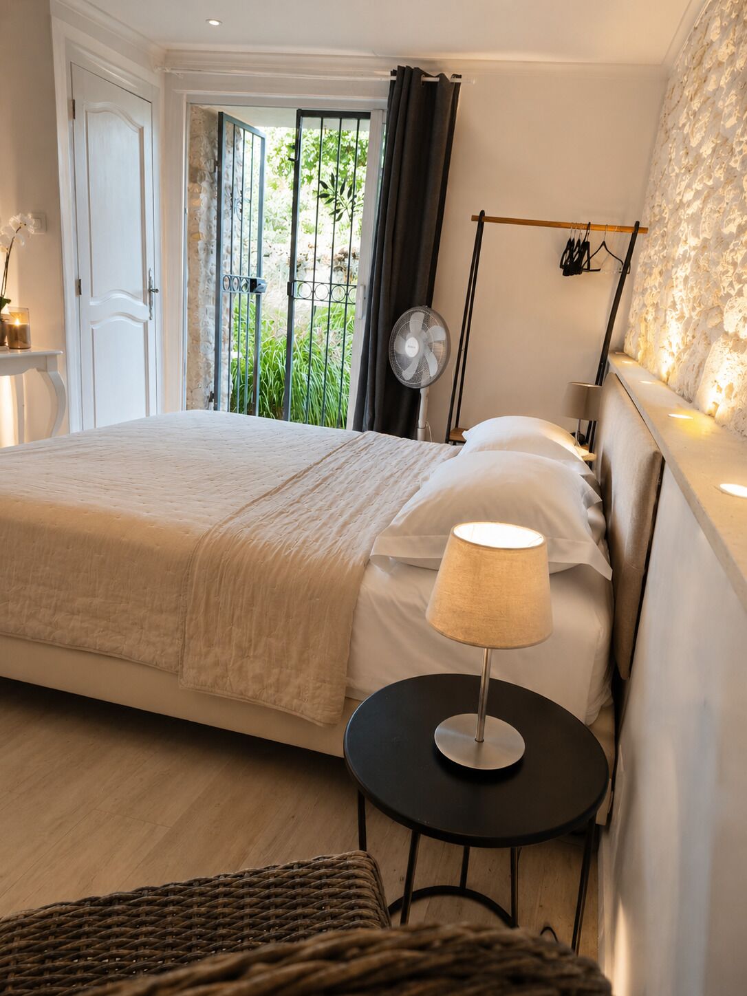 A bedroom with a bed, bedside table, lamp, and a view of greenery outside.
