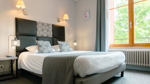 Een net opgemaakt bed met een grijs hoofdbord en gekleurde kussens, een nachtkastje met een lamp, en een raam met uitzicht op groen.