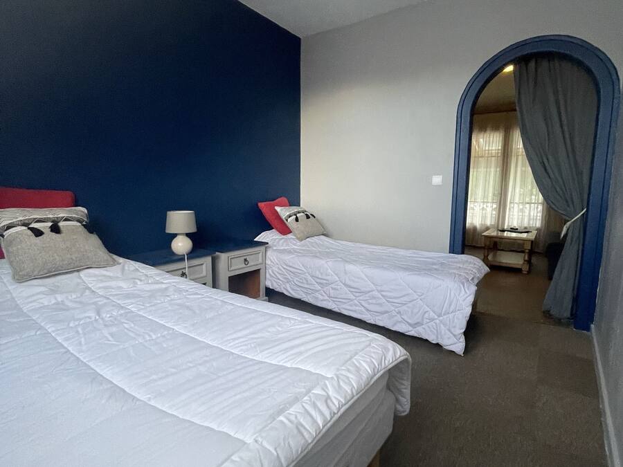 A hotel room with two single beds, a nightstand, a lamp, and a view through an arched doorway.