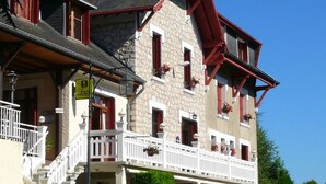 Un immeuble de deux étages avec une façade en pierre, des encadrements de fenêtres rouges et un balcon doté de balustrades blanches.