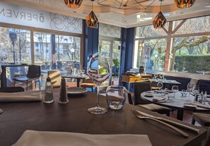 A restaurant interior with tables set for dining, large windows offering a view of the outdoors, and a logo for Logis Hotels. - Hôtel Restaurant Ô Pervenches (Chambery)