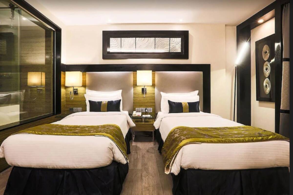 A hotel room with two beds, a large window, a wall-mounted artwork, and a bedside table with a lamp.