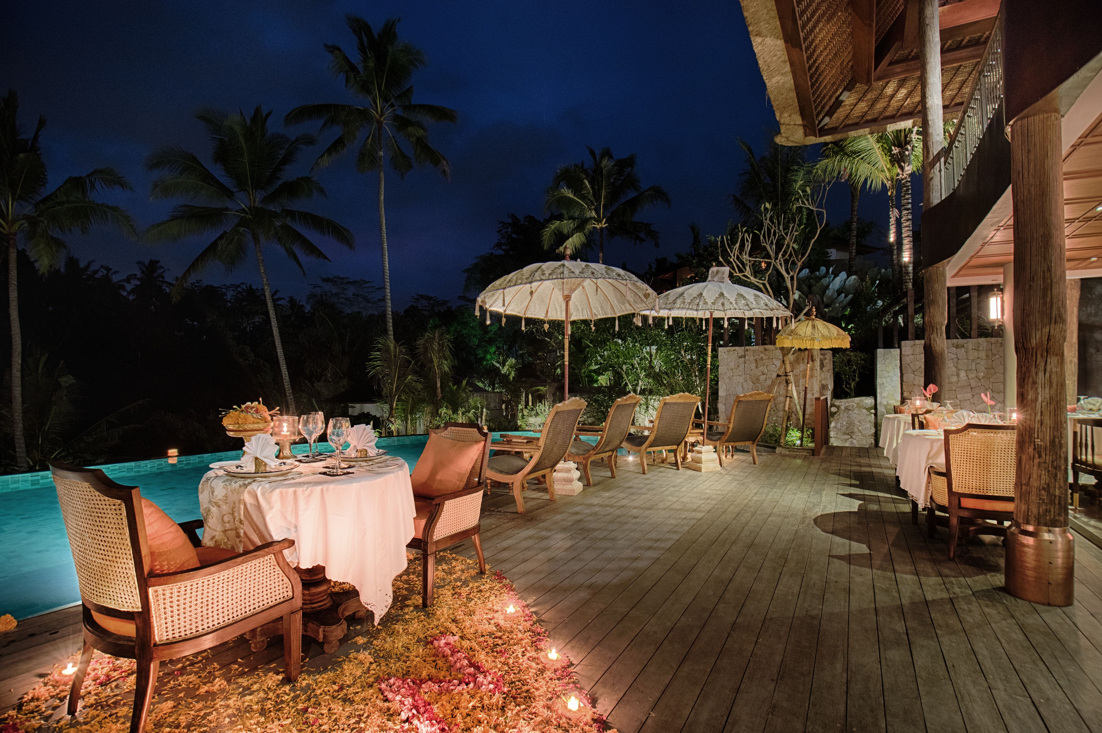 An outdoor dining area with a pool, wooden deck, and palm trees at night.