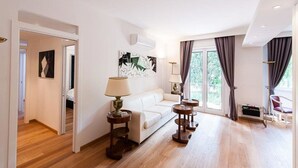 Amplia sala de estar con un sofá blanco, una mesita de centro y una lámpara. Hay dos cuadros en la pared y una ventana con cortinas.