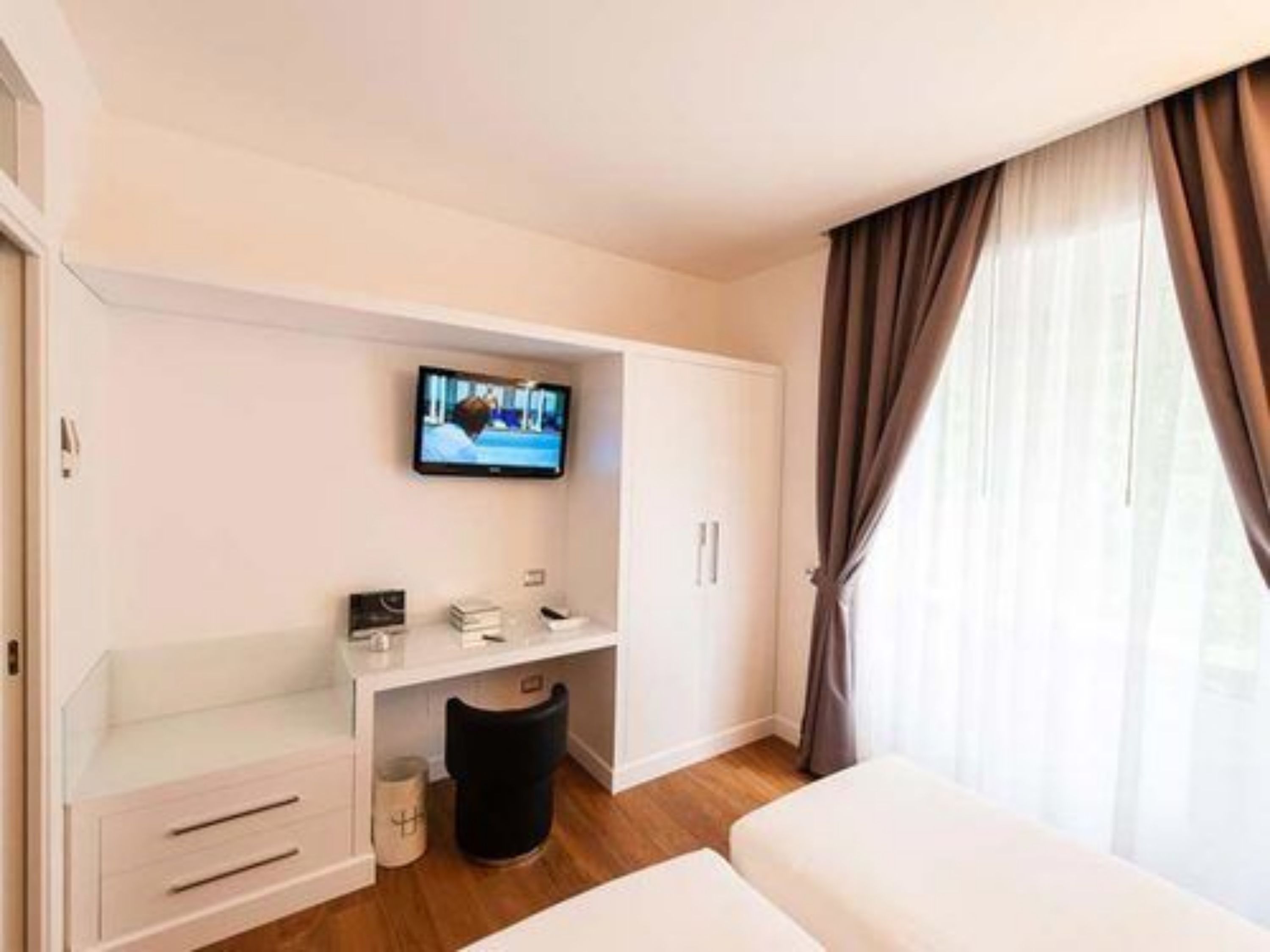 A hotel room with a bed, a TV mounted on the wall, a small desk with a chair, and a window with curtains.