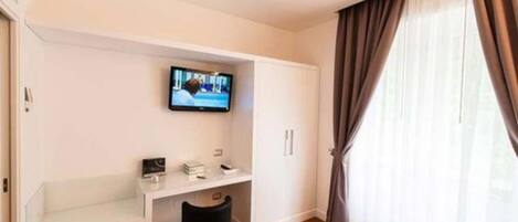 A hotel room with a bed, a TV mounted on the wall, a small desk with a chair, and a window with curtains.