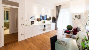 Una sala de estar moderna con una mesa de comedor de vidrio, muebles blancos y un televisor de pantalla plana montado en la pared.