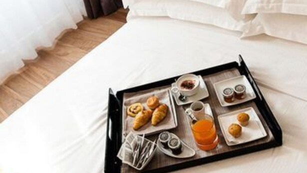 Una cama bien hecha con sábanas blancas, una bandeja con cruasanes, café y jugo, y una mesita de noche con una lámpara.
