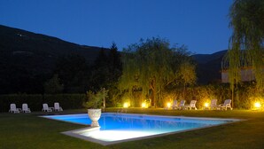 A rectangular swimming pool with a white edge, surrounded by a well-manicured lawn. There are several lounge chairs arranged around the pool, and a large decorative planter is placed in the center.