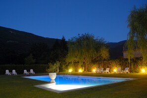 A rectangular swimming pool with a white edge, surrounded by a well-manicured lawn. There are several lounge chairs arranged around the pool, and a large decorative planter is placed in the center.