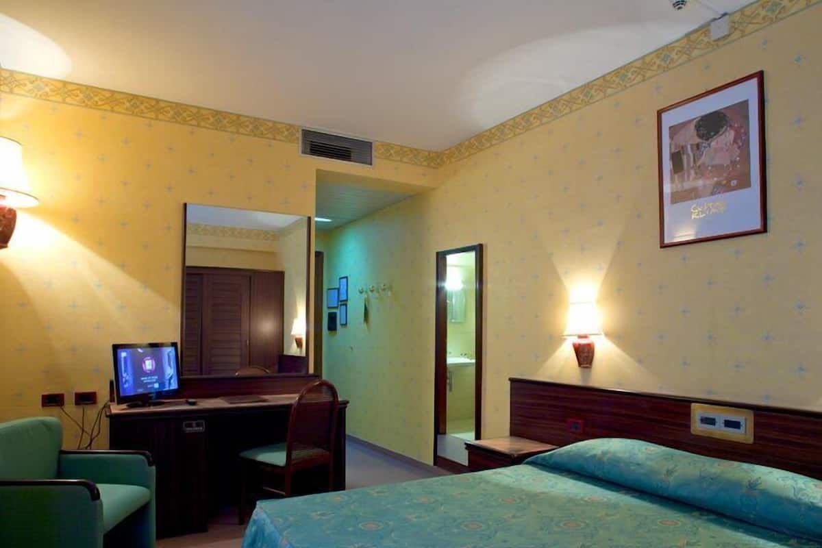 Ein Hotelzimmer mit Bett, Schreibtisch und Stuhl. Auf dem Schreibtisch steht ein Fernseher und an der Wand ein Spiegel.