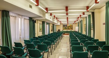Un salón de conferencias con filas de sillas verdes, una larga mesa al frente y cortinas verdes en las ventanas.