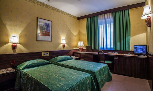 Habitación de hotel con dos camas, un escritorio, una silla, un televisor y una ventana con cortinas verdes.